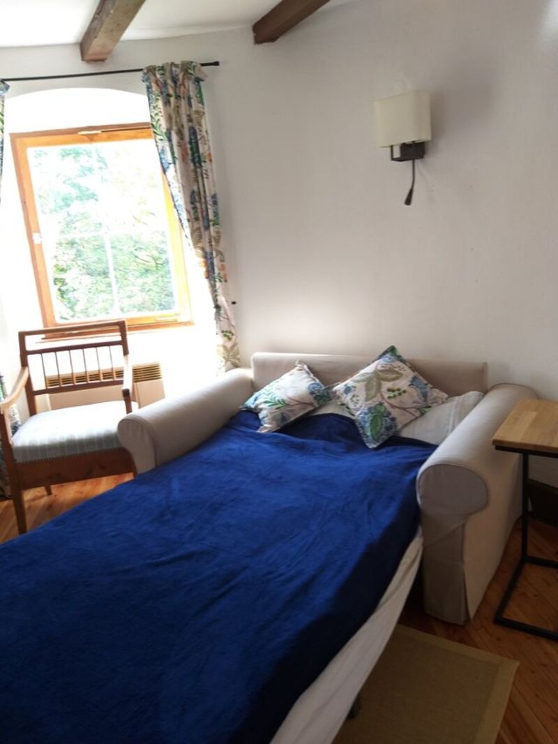 Schlafsofa mit blauer Decke und Blumenkissen vor hellem Bogenfenster mit Blick ins Grüne