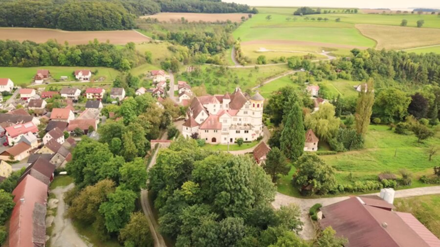 Luftaufnahme von Schloss Laibach: die Gebäude der Vierflügelanlage mit ihren Türmen, eingebettet zwischen Wald, Wiesen und Feldern, im Hintergrund das Dorf